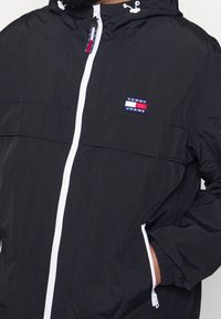 Czarna lekka kurtka z kapturem, z białym suwakiem, elastycznymi mankietami i widoczną naszywką z logo na piersi. Gładka tekstura.