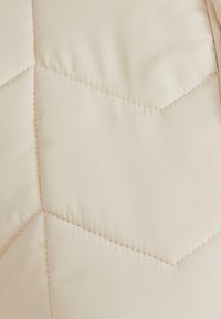 Tessuto trapuntato in un morbido beige, caratterizzato da un motivo a zigzag con cuciture visibili, creando un aspetto liscio e testurizzato.