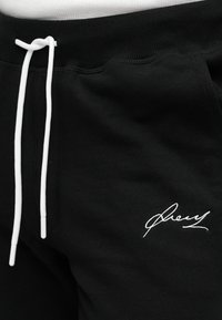 Μαύρο παντελόνι φόρμας με rib ύφασμα στη μέση και λευκά κορδόνια. Διαθέτει λευκό κεντητό κείμενο κοντά στο τελείωμα, που αναδεικνύει χαρακτηριστικό στυλ.