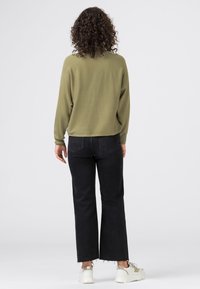 Person with curly hair stands facing away, wearing an olive green sweater, black straight-cut jeans, and white sneakers on a plain background.