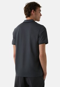 Tmavě šedá polo tričko s krátkým rukávem, vyrobené z hladkého materiálu. Obsahuje klasický límec a decentní branding na zadní straně.