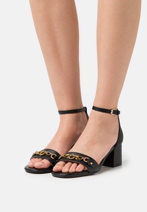 Sandali neri in pelle con tacco a blocco e cinturino alla caviglia, dotati di un'accativo in catena dorata sulla zona delle dita, design open-toe e superficie liscia.