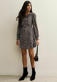 Zwarte en beige luipaardprint jurk met lange mouwen en een gerimpelde taille, gecombineerd met zwarte enkellaarsjes en een kleine zwarte handtas.