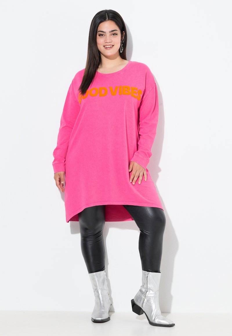 Vestito felpa rosa con stampa arancione "GOOD VIBES", maniche lunghe, orlo asimmetrico, abbinato a leggings neri e stivali alla caviglia argento.