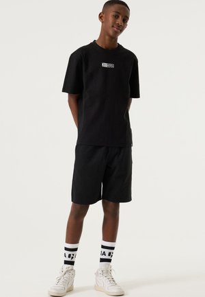 Giovane in piedi, indossa una maglietta nera con la scritta "Energetic Rewind", pantaloncini neri, sneakers bianche alte e calzini bianchi con strisce nere e testo.