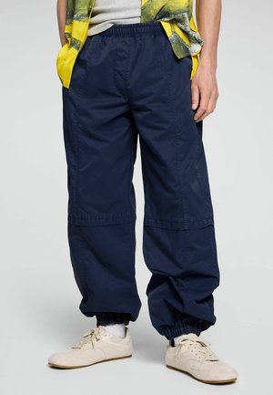Person trägt marineblaue Jogginghose mit elastischen Bündchen, beige Sneaker, weiße Socken, ein gelb-grünes offenes Hemd und ein graues Top darunter.