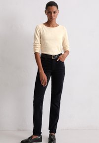 T-shirt crème à manches longues avec un décolleté bateau, associé à un pantalon en velours côtelé noir et une ceinture noire, agrémenté de mocassins noirs brillants.