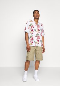 Kurzarm, weißes Blumenshirt mit roten und gelben Mustern, kombiniert mit beigen Cargo-Shorts und weißen Sneakers.