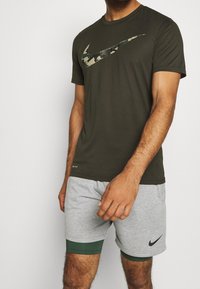 Camiseta Nike verde oliva con un logo de camuflaje, combinada con pantalones cortos grises que tienen un acento verde y un swoosh negro de Nike.