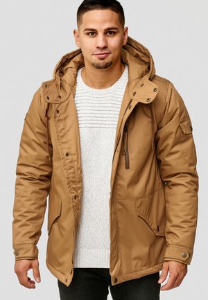 Parka isolante marron avec capuche, fermeture éclair à l'avant, poches latérales et poignets élastiques, porté sur un pull léger texturé et un jean foncé.