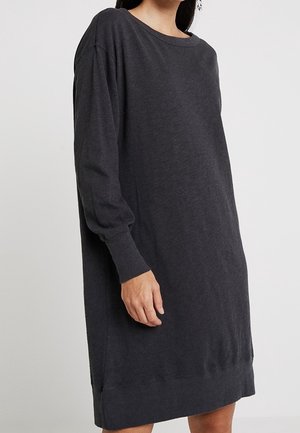 Femme portant une robe ample en tricot gris foncé à manches longues, debout devant un fond clair uni.