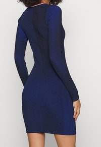 Robe en tricot bleu marine avec un design texturé à côtes, manches longues et silhouette ajustée. Comprend un détail de décolleté rouge contrastant.
