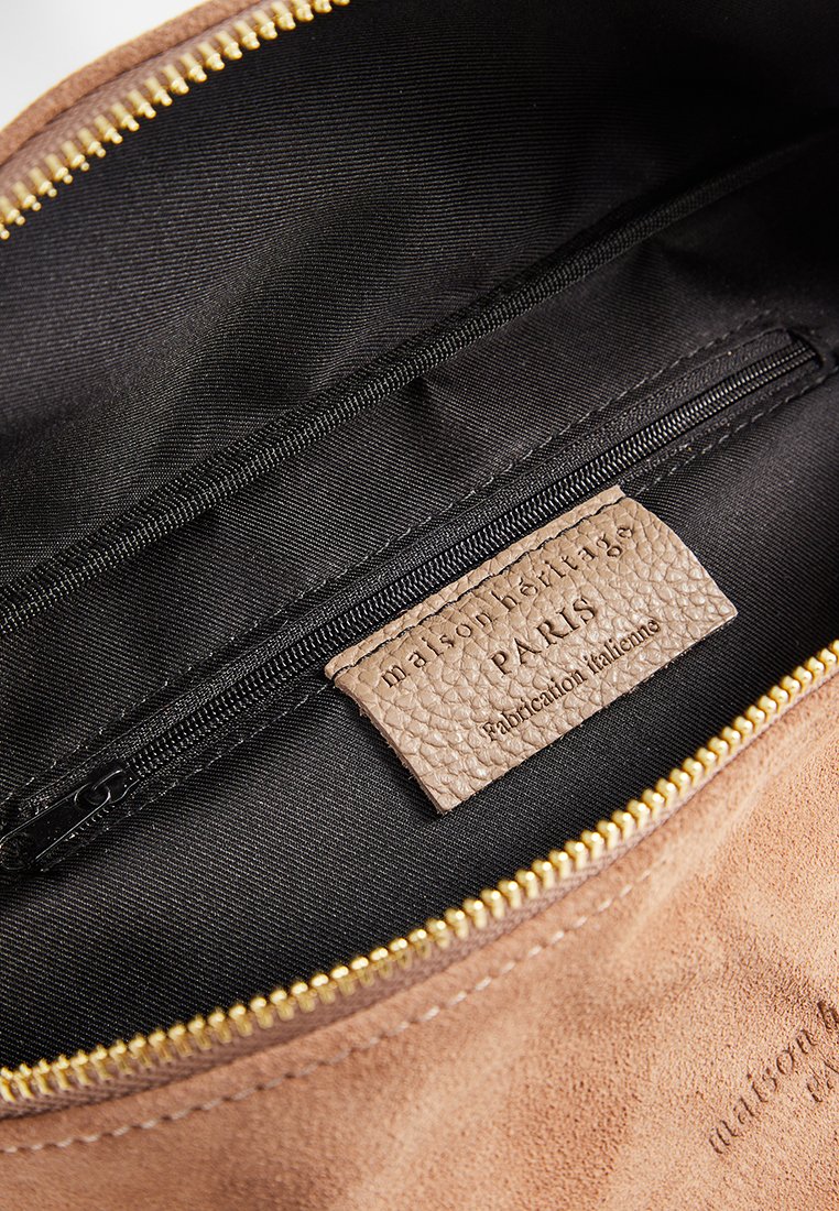 Intérieur d'un sac en daim marron avec une doublure noire, une fermeture éclair dorée et une étiquette en cuir beige avec du texte en relief. Inclut une poche zippée.