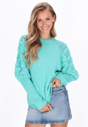 Turquoise gebreide trui met gestructureerde kabelstrikmouwen, geribbelde manchetten en een ronde halslijn, gecombineerd met een denim minirok.
