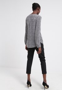 Vrouw in een wit en zwart gestreept blouse met lange mouwen, zwarte cropped pants en zwarte hoge hakken, staand met haar rug zichtbaar.