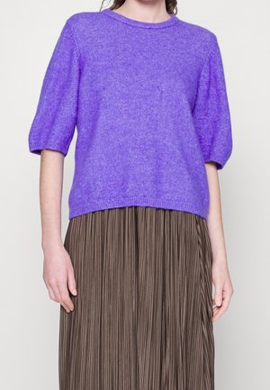 Frau trägt einen lila kurzärmligen Pullover zu einem braunen plissierten Rock, sie steht vor einem schlichten, hellen Hintergrund.
