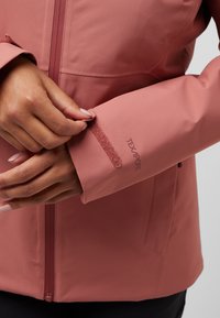 Waterdichte roze jas van textuur materiaal. Voorzien van een ritssluiting aan de voorkant en verstelbare cuffs met klittenband. Het logo leest "TEXAPORE."