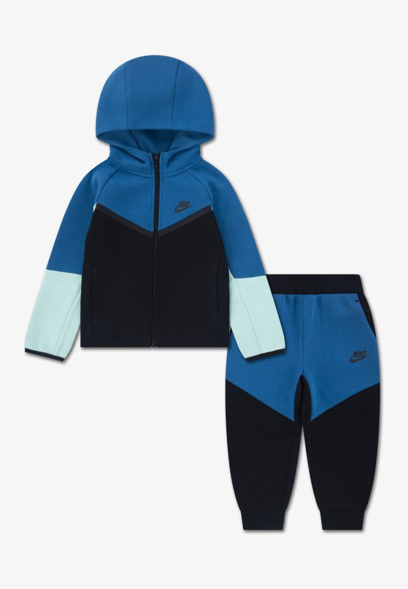 Zila Nike sporta tērps, kas sastāv no džemperim ar kapuci ar rāvējslēdzēju un melniem akcentiem, kā arī atbilstošiem biksēm, izgatavots no mīkstas auduma ar krāsu bloku dizainu.