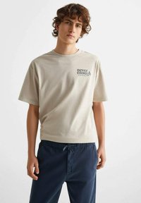 Beige bomulls t-shirt med korta ärmar, med en tryckt "never enough" design i grått på vänster sida. Matchad med marinblå drawstring-byxor.