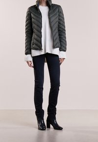 Kvinde iført en mørkegrøn, quiltet jakke over en hvid sweater, sorte skinny bukser og sorte hælede ankelstøvler, der står på et simpelt gulv.