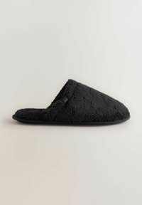 Svart toffel med en texturerad, mjuk ovandel i tyg och en rundad tåkonstruktion. Har en platt svart sula för inomhusbruk.