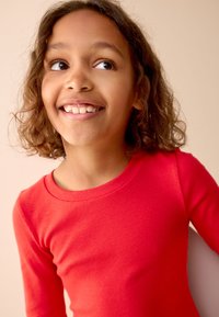 Red ribbed long-sleeve top with a rounded neckline. Features a soft texture and a fitted shape. Background in neutral tones.