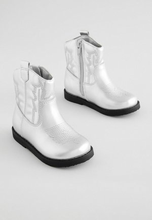 Stříbrné kovbojské boty s lesklým povrchem, zdobené vzorovaným stehem a bočním zipem, usazené na černé podrážce pro kontrast.