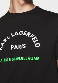 Černé bavlněné tričko s bílým a neonově zeleným potiskem: "KARL LAGERFELD PARIS" a "21 RUE ST-GUILLAUME," s kulatým výstřihem.