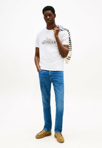 Camiseta blanca de algodón con un logo azul y dorado, combinada con jeans de mezclilla azul claro y zapatos marrones. Un sudadera a rayas está colocada sobre un hombro.