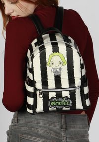 Mochila a rayas en blanco y negro con un diseño de personaje de dibujos animados, que cuenta con un bolsillo frontal con cremallera y un logo de "Beetlejuice". Textura suave.
