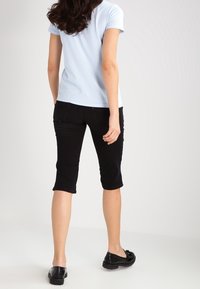 Svarta capribyxor med smal passform, tillsammans med en ljusblå kortärmad skjorta. Skorna är svarta lägenheter. Bakgrunden är enfärgad.