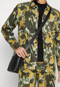 Kamouflage-mönstrad outfit i gröna, gula och blå nyanser, med en svart lädercrossbody-väska med en ficka på framsidan och rem.