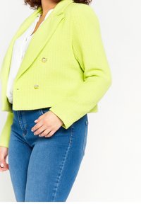 Limetkově zelený zkrácený blazer s strukturovanou tkaninou, dvouřadým zapínáním a stříbrnými knoflíky, spárovaný s modrými džíny s vysokým pasem.