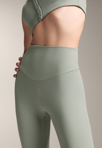 Csíkozott, magas derekú leggings, tompa zöld színben, varratmentes kialakítással és sima textúrával. Diszkrét gombdísz a tetején.