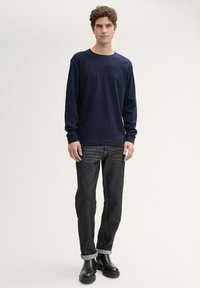 Marineblaues Langarmhemd aus Baumwollstoff, Rundhalsausschnitt, lockere Passform; kombiniert mit dunkelblauen Jeans und schwarzen Knöchelstiefeln mit dicker Sohle.