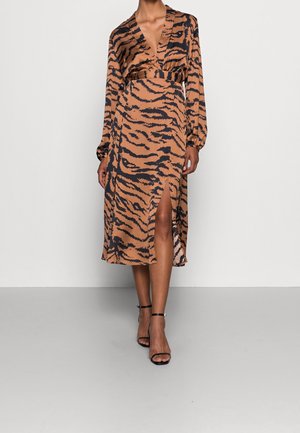 Vrouw draagt een midi-jurk met lange mouwen in bruin en zwart tijgerprint, met een split aan de voorkant en zwarte enkellaarzen met riempjes en hoge hakken.