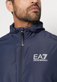 Navy windbreaker with a zipper and adjustable hood. Features EA7 logo in white on the front. Smooth, lightweight material with minimal texture.