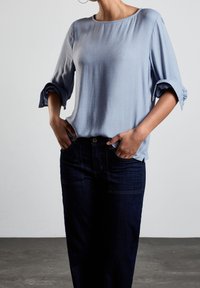 Ljusblå, mjuk tygblus med trekvartslånga ärmar och rullade manschetter, matchad med mörka jeans i denim med framfickor.