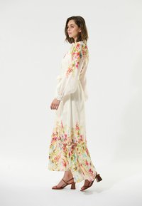 Fließendes weißes Maxikleid mit bunten Blumenmustern an den Ärmeln und am Saum, mit elastischen Manschetten und einem gebundenen Taillengürtel. Getragen mit braunen Blockabsatzsandalen.