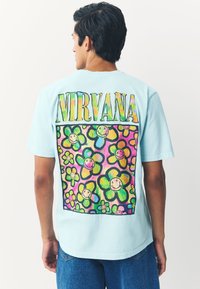 Hellblaues T-Shirt mit einem großen, bunten Blumenmotiv auf der Rückseite, das lächelnde Gesichter zeigt, und dem Wort "NIRVANA" darüber.