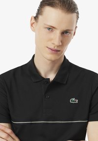 Camiseta polo negra con una tapeta de dos botones, que presenta un logo de cocodrilo verde y un acento de rayas horizontales en verde y blanco. Textura de tela suave.
