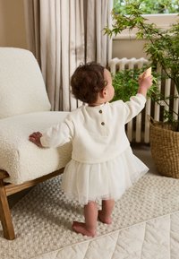 Weißer Pullover mit Knöpfen auf der Rückseite und ein durchsichtiger weißer Tüllrock. Kind steht neben einem strukturierten beigen Stuhl und greift nach einer grünen Pflanze.