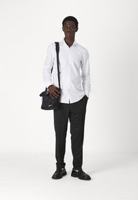 Chemise blanche à manches longues, pantalon foncé à motifs, chaussures en cuir noires et petit sac en bandoulière noir. Design simple et coupe structurée.