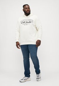 Cremefarbener Hoodie mit aufgedrucktem Logo, kombiniert mit blauen Jeans und weißen Sneaker. Der Hoodie hat eine Kordel und gerippte Bündchen.