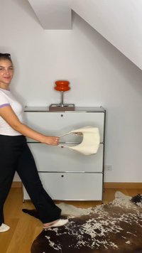Krems krāsas soma ar izliekta forma un plāniem rokturiem, turēta pret minimālisma skapja un dekoratīvās lampas fonu.