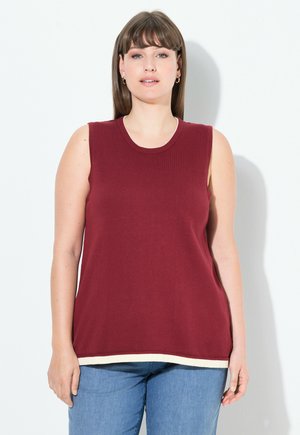 Femme aux cheveux bruns raides portant des boucles d'oreilles en or, un haut sans manches bordeaux avec des bordures blanches et un jean bleu, se tenant contre un fond blanc.