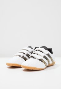 Zapatillas deportivas blancas con tres bandas negras y doradas, que presentan un upper texturizado y una suela de goma. Cerraduras de velcro.