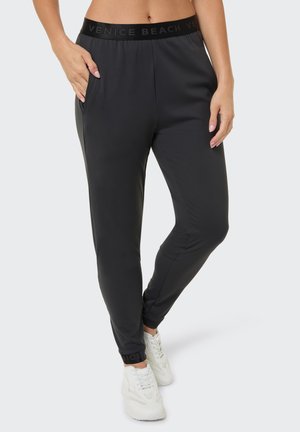 Woman wearing black tapered pants with "Venice Beach" waistband and white sneakers, standing with one hand in pocket.