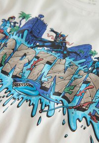 T-shirt bianca in cotone con una stampa grafica colorata che presenta la scritta audace "MARTINI", circondata da onde blu, personaggi e uno squalo.