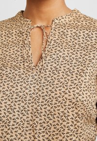 Top beige avec un motif floral composé de petites fleurs noires et orange. Encolure à ouverture clé avec un lien de fermeture. Texture douce.